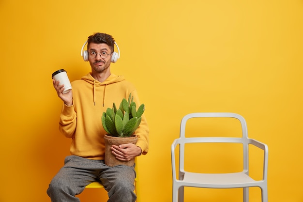 Puzzled confused European man listens favorite music in headphones drinks coffee to go and sits alone near empty chair holds potted cactus