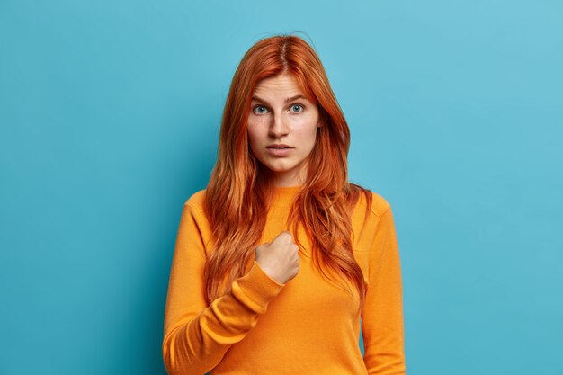 Puzzled confused redhead woman points at herself and asks who me dressed in casual orange jumper cannot understand why she was chosen dressed casually.