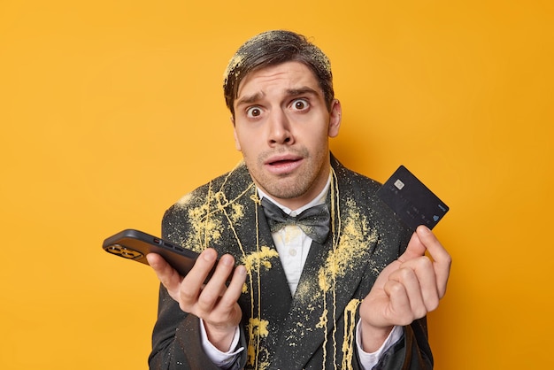 Озадаченный смущенный потрясенный мужчина держит пластиковую банковскую карту, а смартфон не знает, как перевести деньги, носит черную формальную одежду, позирует на желтом фоне, пытается заплатить деньги в ресторане