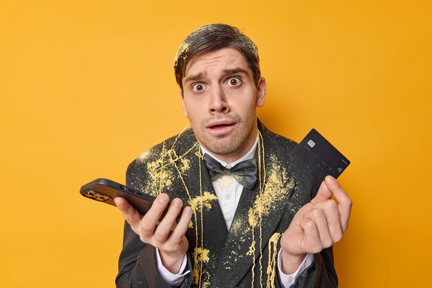 困惑した混乱したショックを受けた男はプラスチックの銀行カードを持っており、スマートフォンはお金を転送する方法を知りません黄色の背景に対して黒いフォーマルな服のポーズを着てレストランでお金を払おうとします