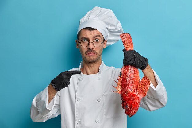 puzzled cook points at big red sea fish, asks advice what to cook from product, needs new recipe, wears white uniform