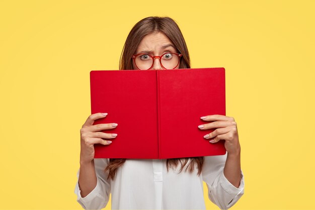 Puzzled dark haired woman raises eyebrows, looks with puzzlement over red textbook, wears white shirt, models against yellow wall, learns new information, prepares home assignment, reads story