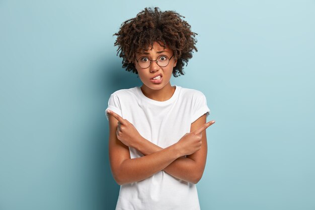 Puzzled dark skinned woman crosses hands over chest, clenches teeth, cannot make choice between two items