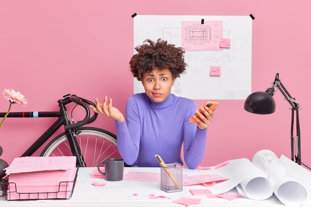 Бесплатное фото Озадаченная темнокожая женщина работает за компьютером, держит мобильный телефон и выражает сомнение, со скептическим выражением лица делает зарисовки