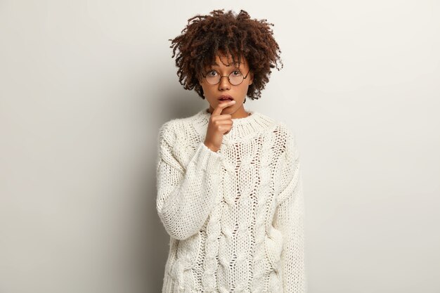 Puzzled dark skinned young female looks nervously, listens important information, opens mouth from amazement, wears round spectacles, white jumper, poses indoor. Human facial expressions concept