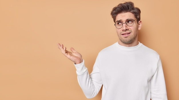 Puzzled displeased adult man has frowned expression demonstrates something umpleased keeps palm raised wears casual white jumper eyeglasses for vision correction isolated over beige studio wall