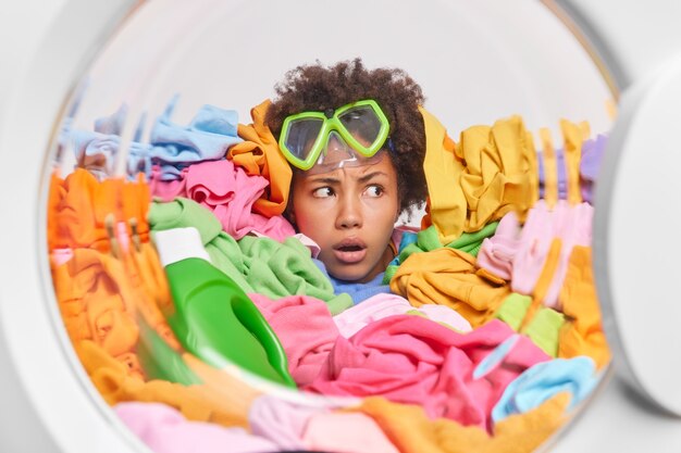 Puzzled displeased Afro American woman looks indignant away wears snorkeling mask drowned in big heap of laundry loads washing machine with clothes has much household duties and responsibilities