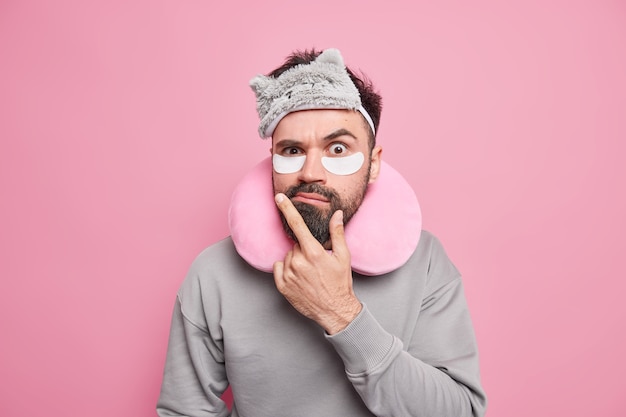 Puzzled displeased bearded man keeps hand on chin doesnt like something wears blindfold on forehead travel pillow around neck casual jumper undergoes beauty procedures applies patches under eyes