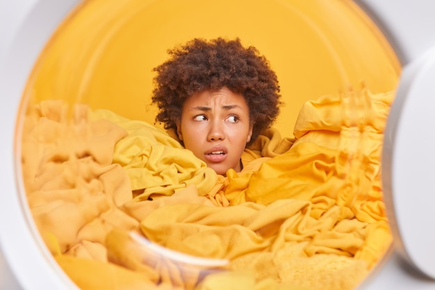 Puzzled displeased curly afro american woman housekeeper looks away drowned in yellow dirty laundry gets out cleaned clothes from washing machine fed up of daily routine and domestic chores