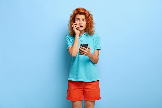 電子メールの確認を受け取らないことにショックを受けた困惑した不機嫌な赤毛の女性は、現代の携帯電話を保持しています