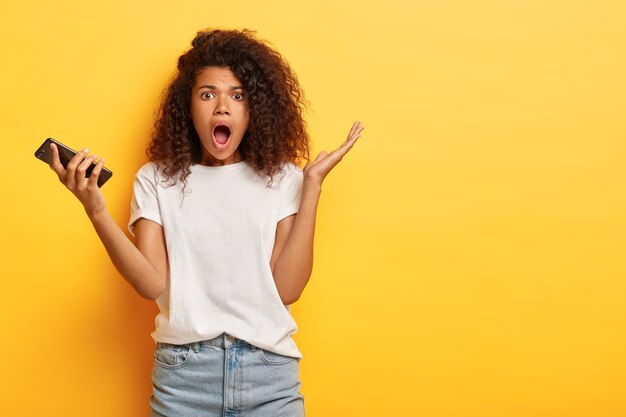 Puzzled emotional dark skinned woman holds smartphone, opens mouth widely,  wears white t shirt, raises palm with bewilderment, isolated on yellow studio wall