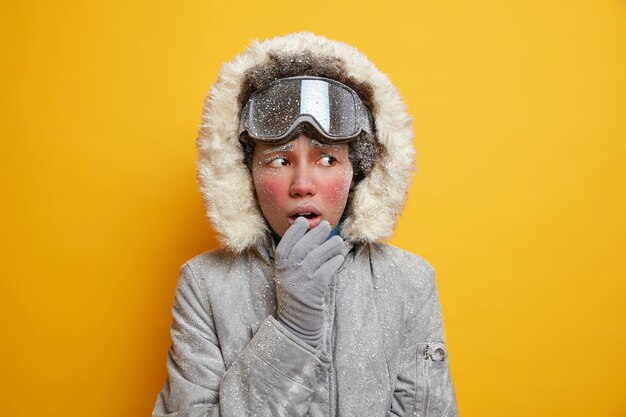 Puzzled ethnic woman with face covered by ice feels cold looks away wears winter jacket gloves freezes during frosty weather.