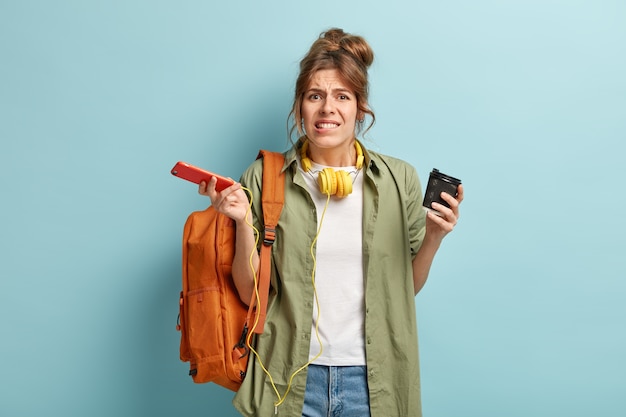 Puzzled female blogger holds mobile phone and takeaway coffee, can not make transaction or payment