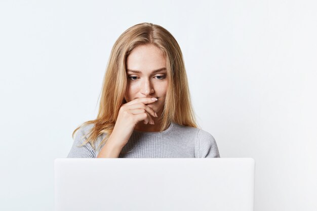 Puzzled female with beautiful appearance reads news online, being focused on laptop computer. Young college student works at his diploma paper or thesis, uses modern technology, isolated on white