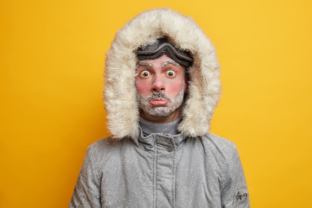 Озадаченный замерзший молодой человек, покрытый снегом, проводит весь день на открытом воздухе в холодную морозную погоду при низких температурах, будучи активным лыжником, одетым в теплую куртку.