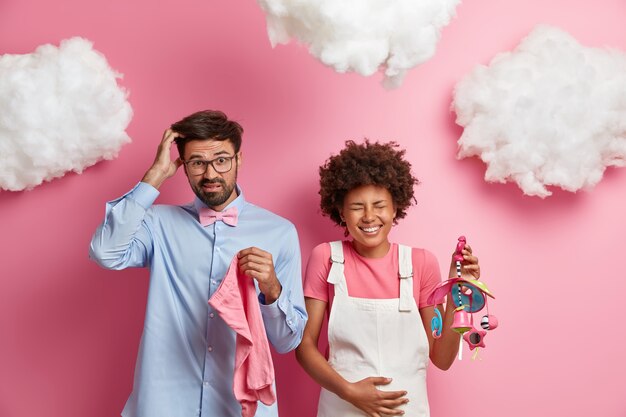 Puzzled future father scratches head as doesnt know what clothes to buy for unborn child, overjoyed pregnant woman touches tummy, holds baby mobile, expresses positive emotions, become parents soon