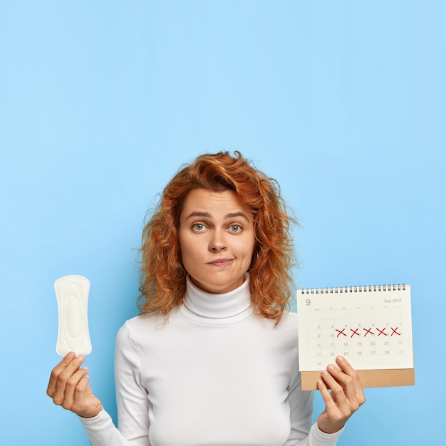 Free photo puzzled ginger woman holds sanitary napkin and menstruation calendar with marked red days