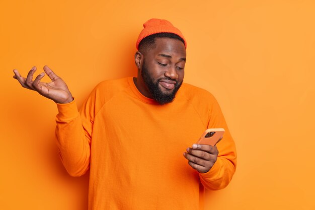 Озадаченный парень поднимает ладони и, сосредоточившись на дисплее смартфона, не может понять, от кого он получил сообщение, носит шляпу и джемпер, изолированные на оранжевой стене