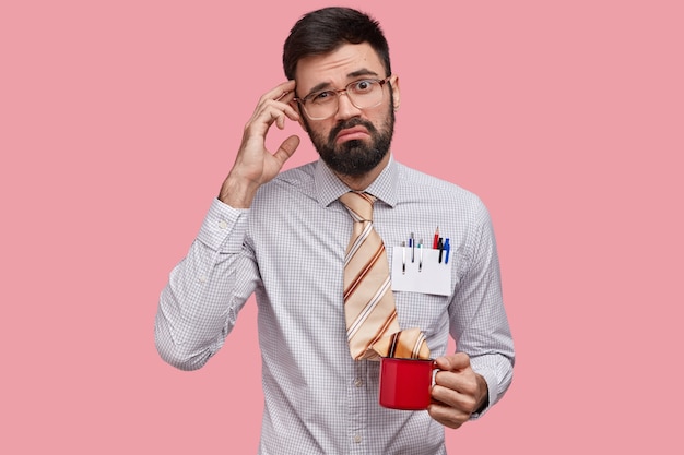 Puzzled heistant unshaven man scratches head, has displeased expression, wears elegant shirt, has tie in cup of drink, has no idea, isolated over pink space