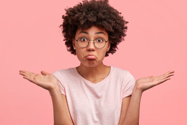 puzzled hesitant african american female with clueless expression, curves lower lip, shrugs shoulders, dressed in casual outfit, poses against pink wall. dark skinned female can`t make decision