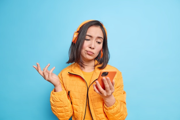  puzzled hesitant Asian woman looks at smartphone display cannot choose song to listen wears wireless headphones on ears wears stylish jacket isolated over blue wall