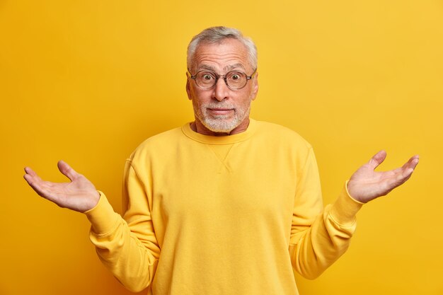 Puzzled hesitant bearded mature man shrugs shoulders in bewilderment spreads hands and looks with uncertainment wears casual sweater isolated over yellow wall makes decision or choice