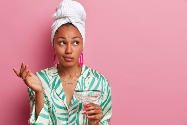 Puzzled hesitant dark skinned healthy woman raises hand and looks doubtfully aside, poses with cocktail