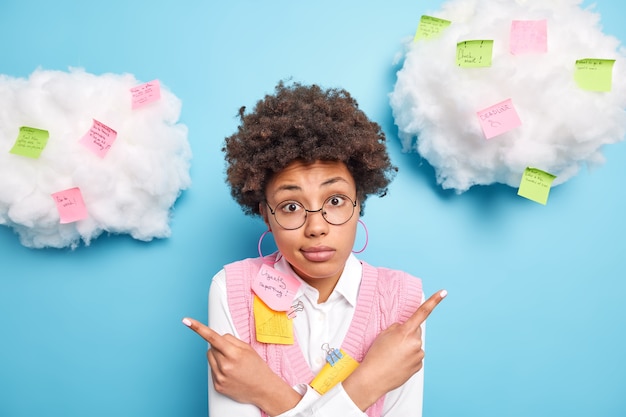 Free photo puzzled hesitant skilled female business worker poses around colorful reminder stickers hesitates between two options wears round spectacles isolated over blue wall