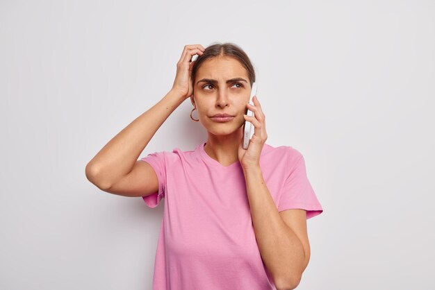 Puzzled hesitant young woman scratches head tries to recall something in mind has telephone conversation dressed casually isolated over white wall