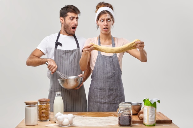 Marito e moglie perplessi guardano con shock l'impasto crudo, preparano la pasticceria, l'uomo frusta gli ingredienti nella ciotola, trascorrono il fine settimana cucinando a casa, posano al banco della cucina. culinario con uova, farina, latte