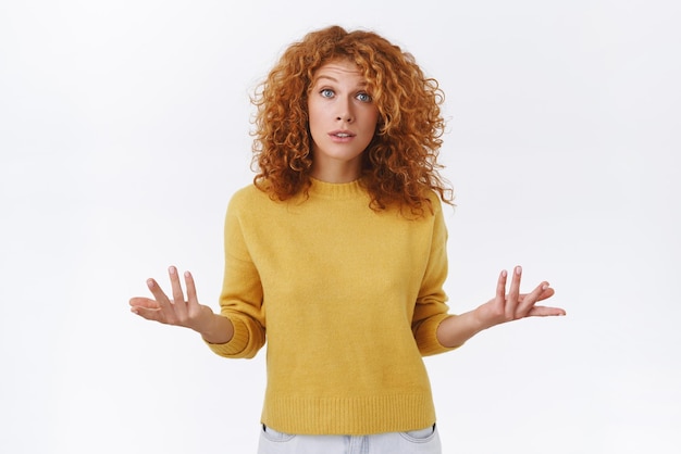 Puzzled indecisive cute redhead curly woman in yellow sweater have misunderstanding raise arms and shrugging troubled lift eyebrows in surprise standing questioned uncertain white background