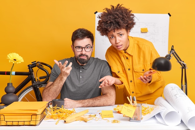 Puzzled interracial woman and man look puzzled while working in office draw sketch of future building construction look indignant pose at desktop concentrated on job 