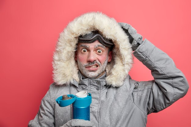 Puzzled male hiker has frozen face shocked by cold conditions in expedition wears warm jacket and ski goggles drinks hot beverage.