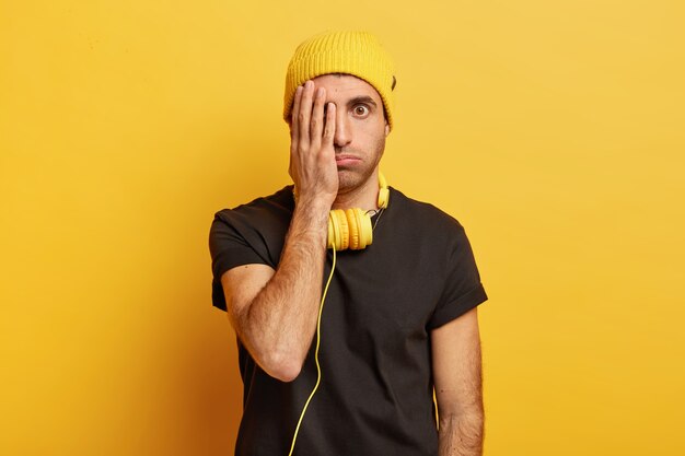 Puzzled male model sighs from tiredness, covers half of face, looks with one eye surprisingly