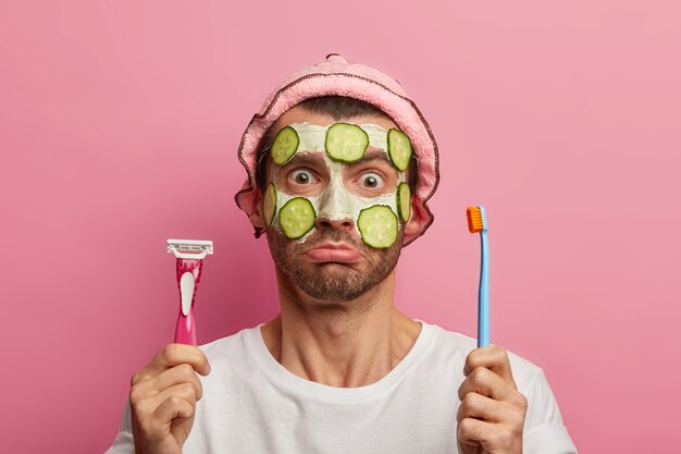 Puzzled man applies nourishing cleansing mask, holds toothbrush and razor, going to shave and brushing teeth, dressed in casual clothes, poses against pink space. Men