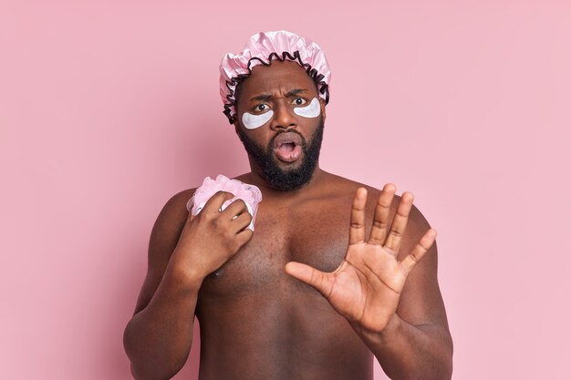 Puzzled man keeps palm forward front takes shower holds sponge wears water proof hat and beauty patches under eyes poses against pink wall