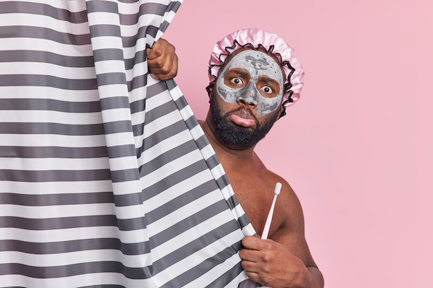 Free photo puzzled man with thick beard applies nourishing clay mask wears bath hat holds toothbrush hides naked body behind shower curtain undergoes daily hygiene procedures isolated on pink wall