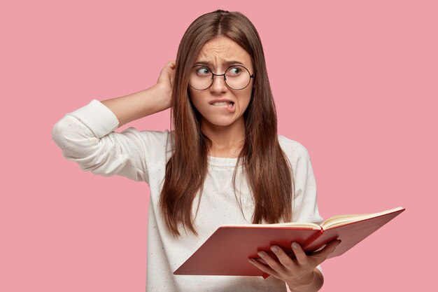 Puzzled nervous woman scratches head, bites lower lip, cant memorize necessary information for seminar, holds opened textbook, dressed in casual white sweater and round spectacles, models indoor