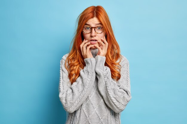 Puzzled nervous woman with long red hair keeps hands on face looks with worried expression afraids of speaking has embarrassed look and reaction on something wears knitted sweater.