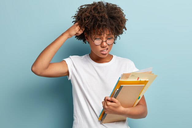 Puzzled questioned pupil scratches head, looks with doubt and displeased expression at papers, carries spiral notebook and book, tries to find answer, dressed in casual white t shirt, isolated on blue wall