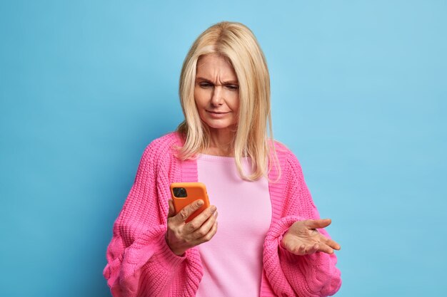 Puzzled retired woman uses mobile phone looks confused smirks face as cannot download new application dressed in casual jumper.