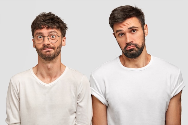 Puzzled sad two bearded young guys stand closely to each other, express negative emotions, look in bewilderment, wear white casual clothes