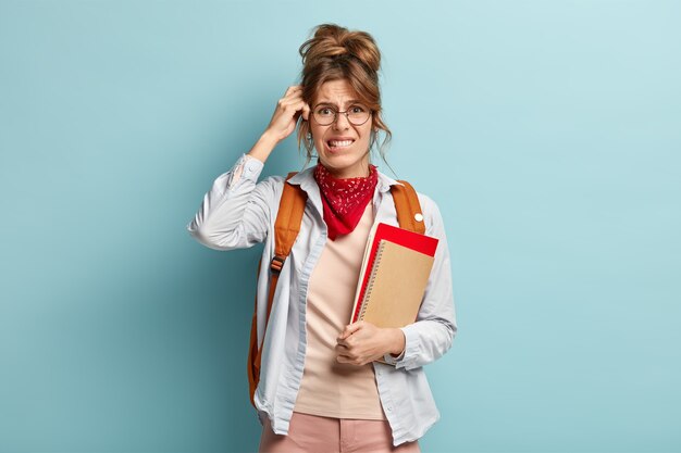 Puzzled schoolgirl holds notebook and spiral diary, bites lips, scratches head as recollects information in mind, wears stylish bandana, transparent glasses, rucksack on back. Studying concept