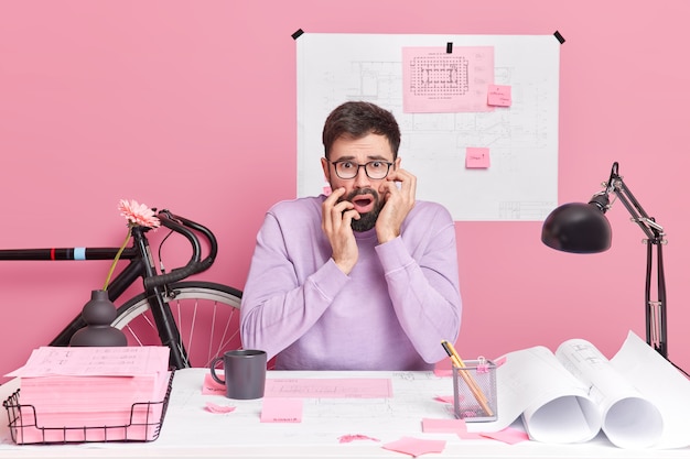 Puzzled shocked bearded man architect works in office with blueprints poses at desktop dressed casually