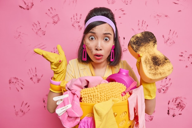 Puzzled shocked brunette young Asian woman wears headband rubber protective gloves stunned to see such mess and chaos in room much dirt holds sponge to reduce dirt stands near laundry basket