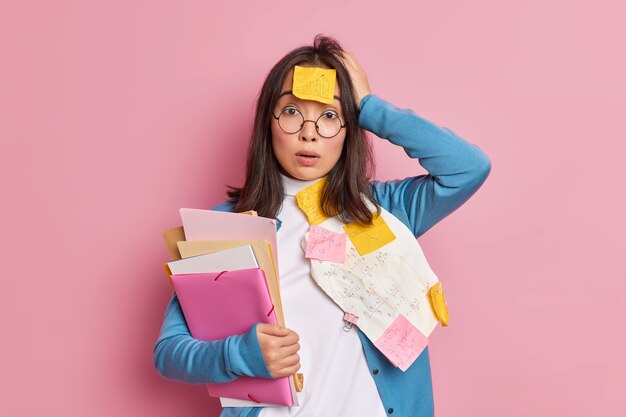 Озадаченная, шокированная женщина-офисная служащая, перегруженная бумагами, ошеломленная тем, что срок окончания исследования, держит папки, держит руку на голове, носит круглые очки.