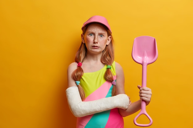 Puzzled small girl in cap and bathing suit, holds toy sand shovel, plays near sea at sandy beach, has bandaged arm after serious injury, isolated on yellow wall. Childhood and summer rest concept