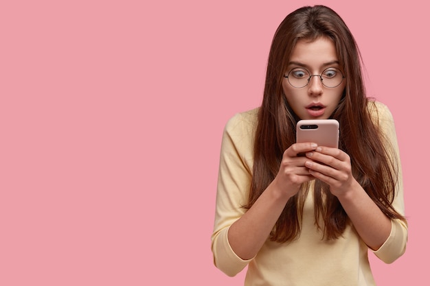 Puzzled stupefied Caucasian woman checks newsfeed in social networks on cell phone, messages with friend, finds out shocking news, stares at screen