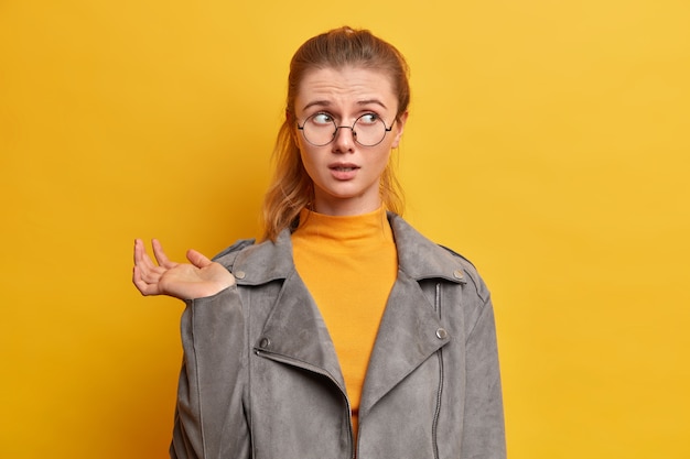 Free photo puzzled surprised caucasian girl looks surprisingly aside, raises hand, has concerned nervous expression, wears optical glasses, raises hand
