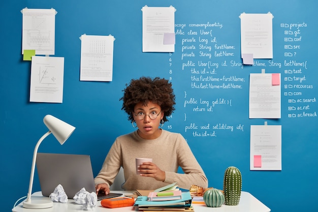 Puzzled surprised computer analyst has coffee break, reads published on web page, wears round spectacles, poses at desktop against blue wall with written information